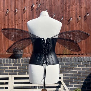 May include: Black leather corset with a lace-up back and black sheer wings. The corset is on a white mannequin.