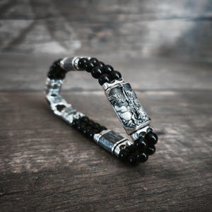 Puede incluir: Una pulsera de doble hebra con cuentas negras pulidas y detalles metálicos rectangulares en tono plateado. Una pieza central plateada destaca con un relieve grabado de una figura alada sosteniendo una espada, junto a espaciadores plateados entre las cuentas.