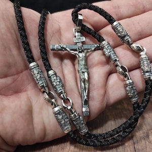 Könnte beinhalten: Ein silberner Kreuzanhänger mit einer detaillierten Darstellung Jesu am Kreuz, der an einer schwarzen geflochtenen Kordelkette hängt. Die Halskette verfügt über verzierte Silberperlen und Verschlüsse und ist ein religiöses Accessoire.