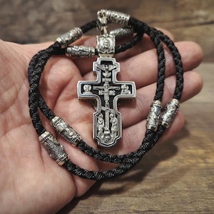 Puede incluir: Un colgante de cruz religiosa plateada con detalles intrincados, suspendido de un cordón trenzado negro. La cruz presenta figuras y símbolos religiosos. Cuentas cilíndricas plateadas están espaciadas a lo largo del cordón.