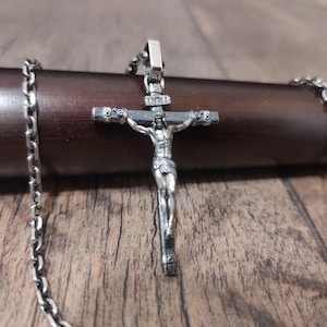 Puede incluir: Un collar de cadena de plata con un colgante de crucifijo de plata detallado que presenta una figura de Jesucristo. El colgante está sobre una superficie de madera.