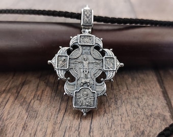 May include: A silver pendant in the shape of a cross with intricate details and multiple small figures. The cross is on a dark brown wooden surface.
