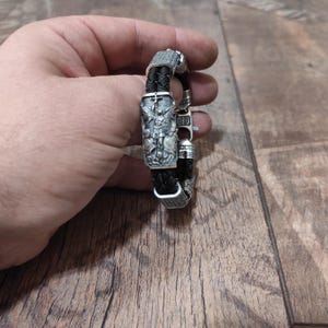 May include: A black leather bracelet with a silver charm featuring a detailed depiction of an angel with a cross. The charm is surrounded by intricate silver accents.