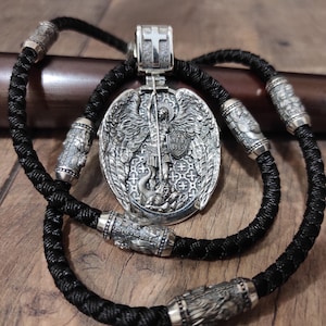May include: A silver pendant necklace featuring a detailed depiction of a saint slaying a dragon. The pendant is oval-shaped with intricate engravings and is suspended from a black braided cord. The necklace includes silver cylindrical beads.