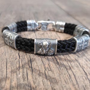 May include: Black braided leather bracelet with silver toned metal beads. The beads feature a Saint Benedict medal and a cross.