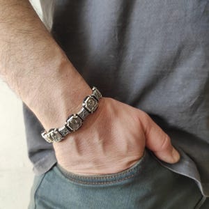 Könnte beinhalten: Ein silbernes Armband mit schwarzen Akzenten und einem sich wiederholenden Muster aus kleinen quadratischen Anhängern. Das Armband wird am Handgelenk getragen.