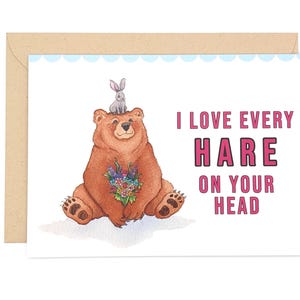 Peut inclure: Une carte de vœux illustrée d'un ours brun tenant un bouquet de fleurs colorées avec un petit lapin gris assis sur sa tête. Le texte sur le côté droit de la carte indique, I LOVE EVERY HARE ON YOUR HEAD, en lettres capitales roses.
