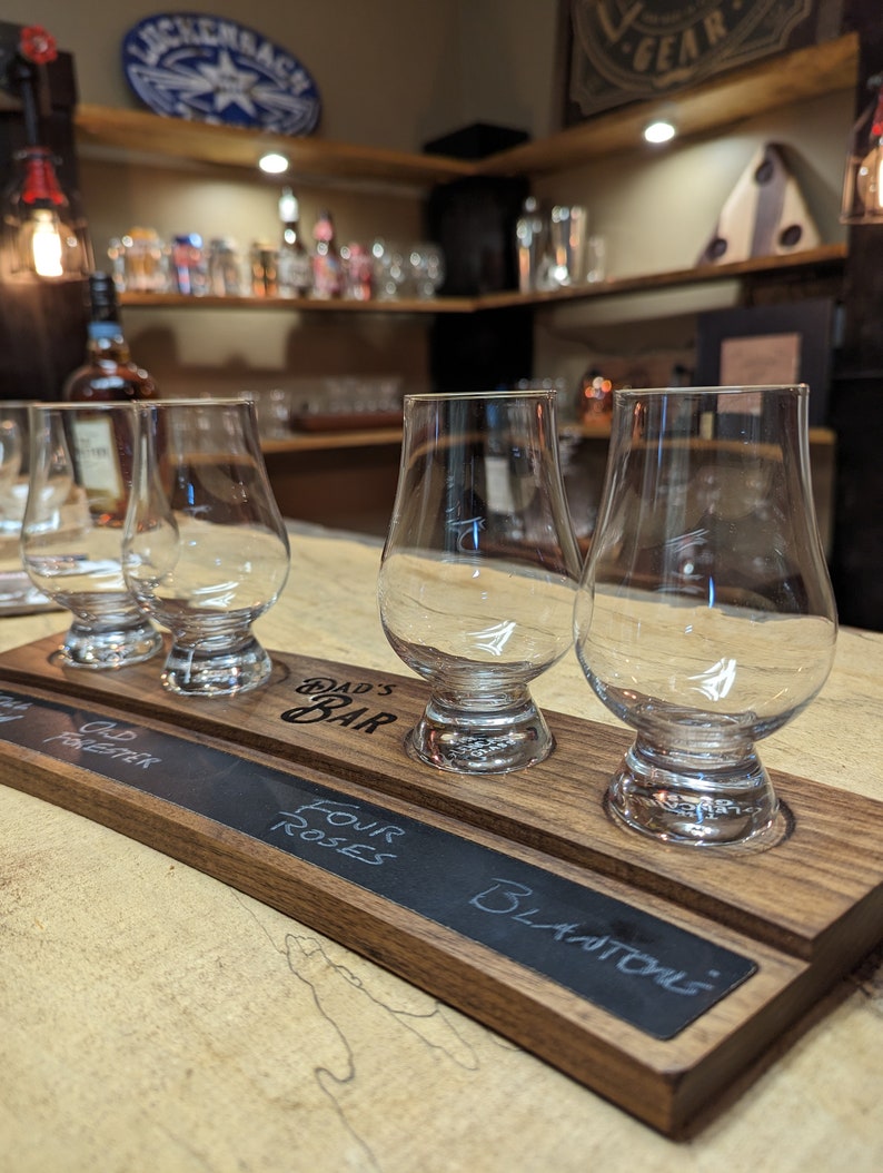 May include: A wooden tasting tray with four glasses and a chalkboard insert. The chalkboard is labeled with the names of different types of whiskey: "Old Forester", "Four Roses", and "Blackstone". The tray is engraved with the words "Dad's Bar".