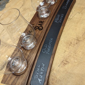 May include: A wooden serving tray with four glass cups and a chalkboard sign. The chalkboard sign has the text "Still Austin", "Old Forester", "Four Roses Blanton's" written on it.