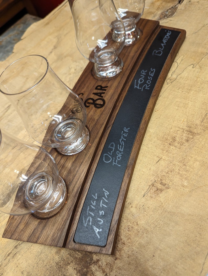 May include: A wooden serving tray with four glass cups and a chalkboard sign. The chalkboard sign has the text "Still Austin", "Old Forester", "Four Roses Blanton's" written on it.