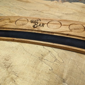 May include: A wooden serving tray with four round indentations for glasses and a long, narrow slot for a bottle. The tray is curved and has the words "Dad's Bar" engraved on the surface.