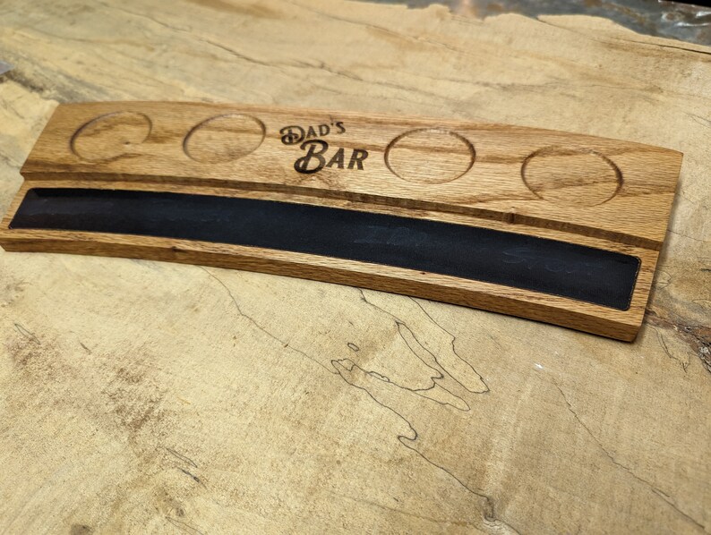 May include: A wooden serving tray with four round indentations for glasses and a long, narrow slot for a bottle. The tray is curved and has the words "Dad's Bar" engraved on the surface.