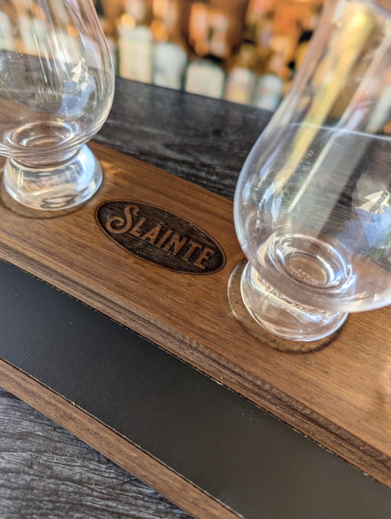May include: A wooden serving tray with a black inlay and the word "Slainte" engraved on it. Two clear glass whiskey glasses are on the tray.