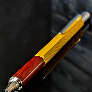 May include: A multi-tool pen with a yellow ruler-marked barrel, a dark wood grip, and a silver tip. The pen's design incorporates a ruler along its side, and a silver clip. The pen is set against a dark, reflective surface.