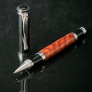 May include: A luxury rollerball pen with a polished burl wood barrel and black accents. The pen features a silver-tone tip and trim, and a matching cap. The cap is detached and placed next to the pen, which is resting on a reflective black surface.