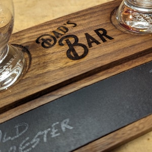 May include: A wooden serving tray with two glass shot glasses and a chalkboard surface. The tray is engraved with the words "Dad's Bar". The chalkboard surface has the word "Old Forester" written on it in white chalk.
