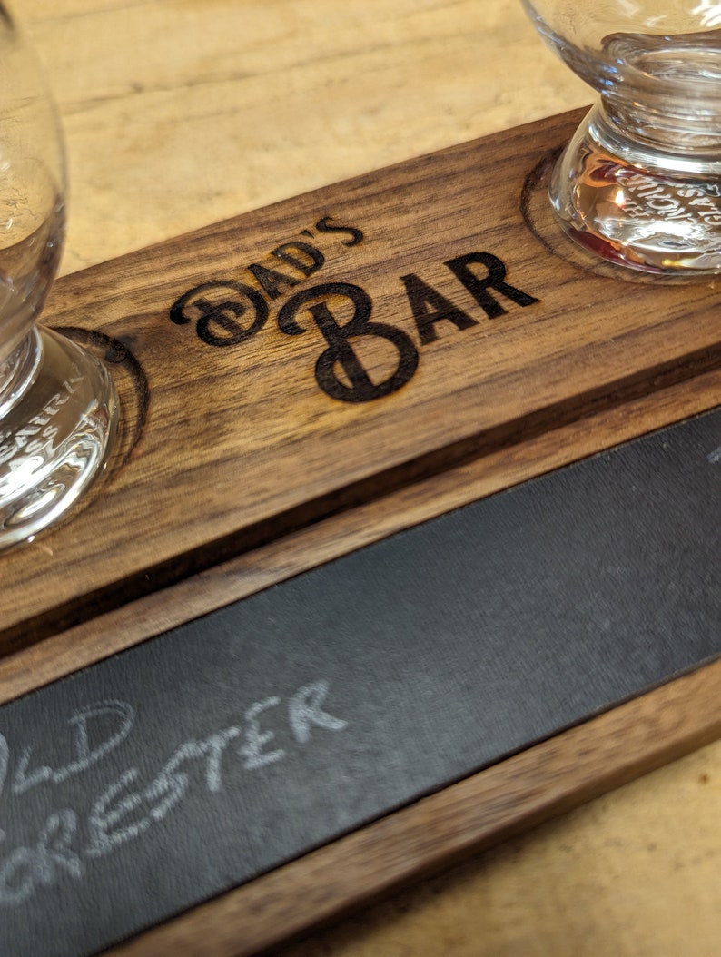 May include: A wooden serving tray with two glass shot glasses and a chalkboard surface. The tray is engraved with the words "Dad's Bar". The chalkboard surface has the word "Old Forester" written on it in white chalk.