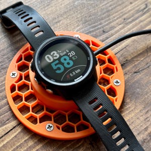 May include: A black Garmin smartwatch with a black band rests on an orange charging base. The watch face displays the time, date, and other metrics. The charging base has a honeycomb design and is secured with screws.