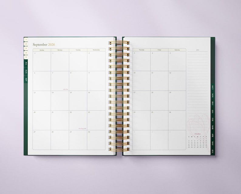 May include: Open planner with a gold spiral binding, displaying the September 2026 calendar. The planner has a dark green cover and tabs, with a small October calendar at the bottom right. The pages are white with a grid layout.