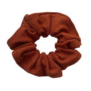 May include: A rust-colored scrunchie made of ribbed fabric. The scrunchie is gathered in a circle and is ready to be worn in hair.