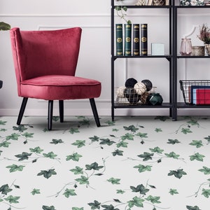 Könnte beinhalten: Ein rosa Samt-Sessel mit schwarzen Beinen steht auf einem weißen Boden mit einem grünen Efeu-Muster. Im Hintergrund befindet sich ein schwarzes Metallregal mit Büchern, Dekoartikeln und einem Korb.