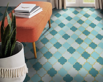 May include: A teal and gold patterned floor tile with a geometric design. The tile is in a room with a light orange ottoman and a green plant in a white pot.