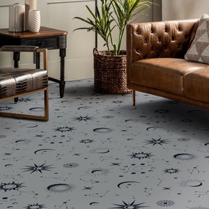 May include: A grey area rug with a celestial pattern featuring moons, stars, and suns in black and white. The rug is shown in a living room setting with a brown leather sofa and a chair.
