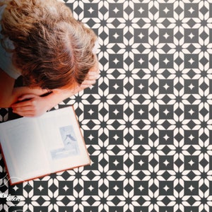Puede incluir: Un patrón de baldosas geométricas en blanco y negro con un diseño de estrella blanca. Una persona está sentada en el suelo leyendo un libro.