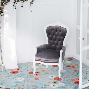 Puede incluir: Un sillón de terciopelo gris con marco blanco se encuentra sobre una alfombra floral azul claro con flores rojas y blancas. El sillón está en una habitación blanca con una chimenea de ladrillo y una puerta blanca con paneles de vidrio.