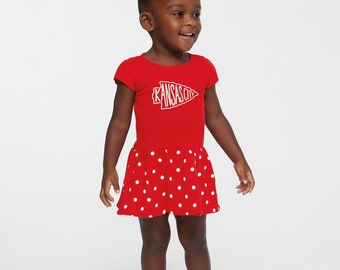 May include: A young child wearing a red dress with white polka dots and a white "Kansas City" logo. The child is also wearing red sneakers. The dress has a short-sleeved top and a flared skirt.