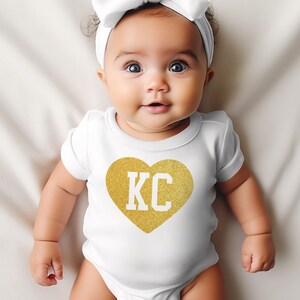 May include: A white baby onesie with a gold glitter heart that says "KC" on it. The baby is wearing a white headband with a bow.