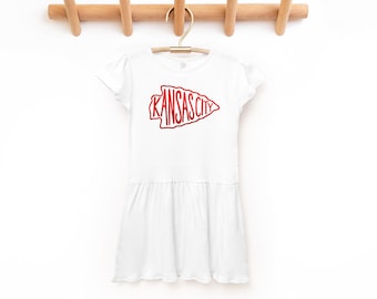 May include: White dress with a red arrow graphic that says "Kansas City" on it. The dress is hanging on a wooden rack.