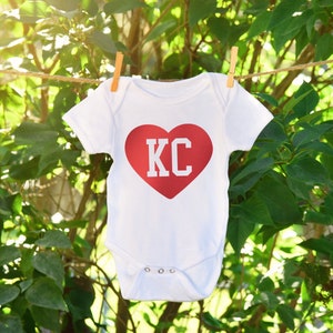 a white onesie with a red heart with KC cut out hanging on a clothes line