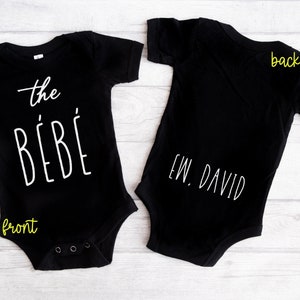 以下が含まれることがあります： 黒のベビーロンパースで、白い文字で「the bébé」と書いてあり、後ろには「EW. DAVID」と書いてあります。