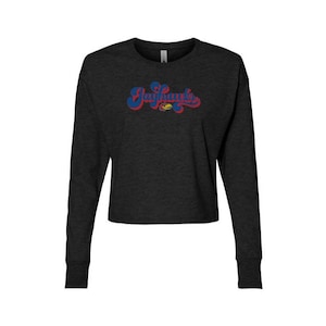 May include: A charcoal grey long-sleeve cropped shirt featuring the word Jayhawks in a blue and red retro-style script font across the chest, with a small yellow and blue bird mascot graphic integrated into the lettering design.