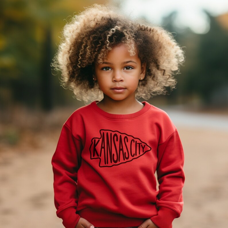 May include: A young person wearing a red sweatshirt with a black graphic of an arrowhead with the text "KANSAS CITY" inside. The person has curly brown hair.
