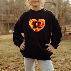 May include: Black sweatshirt with a tie-dye heart design featuring the letters "KC" in black.