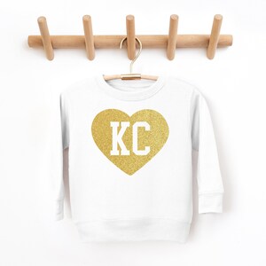 Peut inclure: Sweat-shirt blanc avec un c&oelig;ur en paillettes dor&eacute;es qui dit "KC" en lettres blanches.