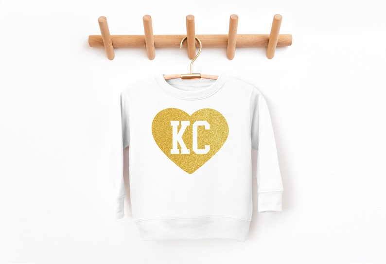 Peut inclure: Sweat-shirt blanc avec un c&oelig;ur en paillettes dor&eacute;es qui dit "KC" en lettres blanches.