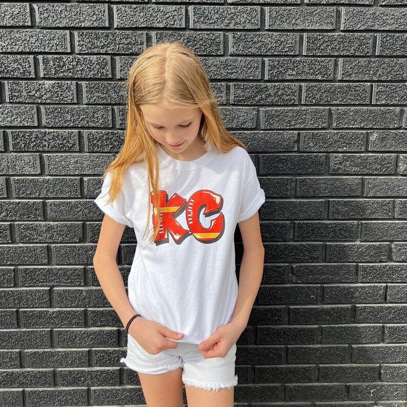 May include: White t-shirt with a red and yellow graphic of the letters "KC".