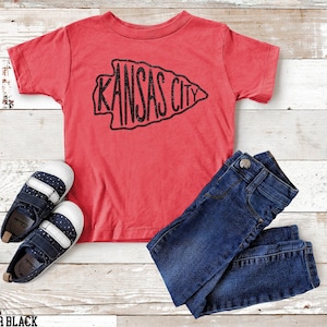 May include: Red short-sleeved t-shirt with a black glitter graphic of an arrowhead with the text "KANSAS CITY" inside. The shirt is laid flat on a woodgrain surface with a pair of blue and white sneakers and blue jeans.
