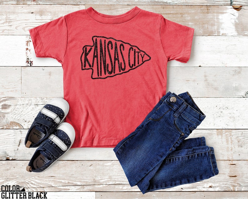 May include: Red short-sleeved t-shirt with a black glitter graphic of an arrowhead with the text "KANSAS CITY" inside. The shirt is laid flat on a woodgrain surface with a pair of blue and white sneakers and blue jeans.