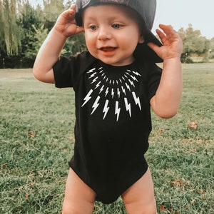 May include: A baby wearing a black bodysuit with a white lightning bolt pattern and a black mesh baseball cap. The baby is standing on a green grassy lawn.