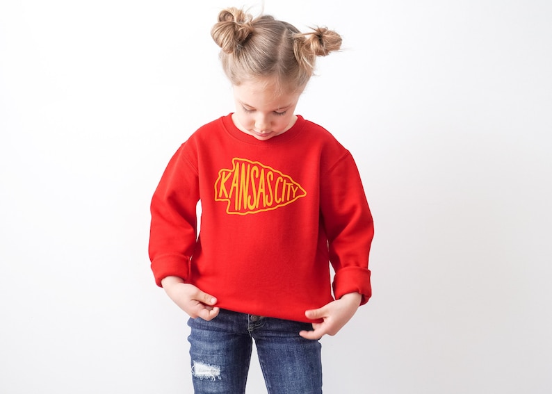 May include: A red sweatshirt with a yellow outline of an arrowhead with the words "Kansas City" inside. A young girl is wearing the sweatshirt.