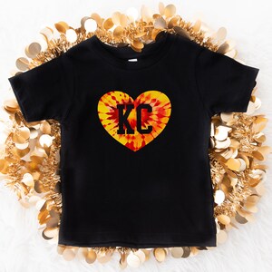 May include: Black t-shirt with a tie-dye heart design and the letters "KC" in black.