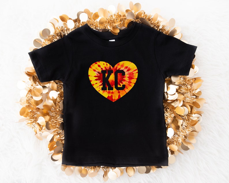 May include: Black t-shirt with a tie-dye heart design and the letters "KC" in black.