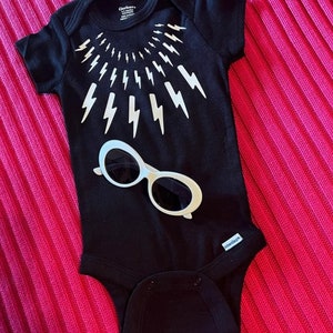 May include: Black baby onesie with a white lightning bolt design around the neck and a pair of white sunglasses on the front. The onesie has the Gerber brand name on the tag.