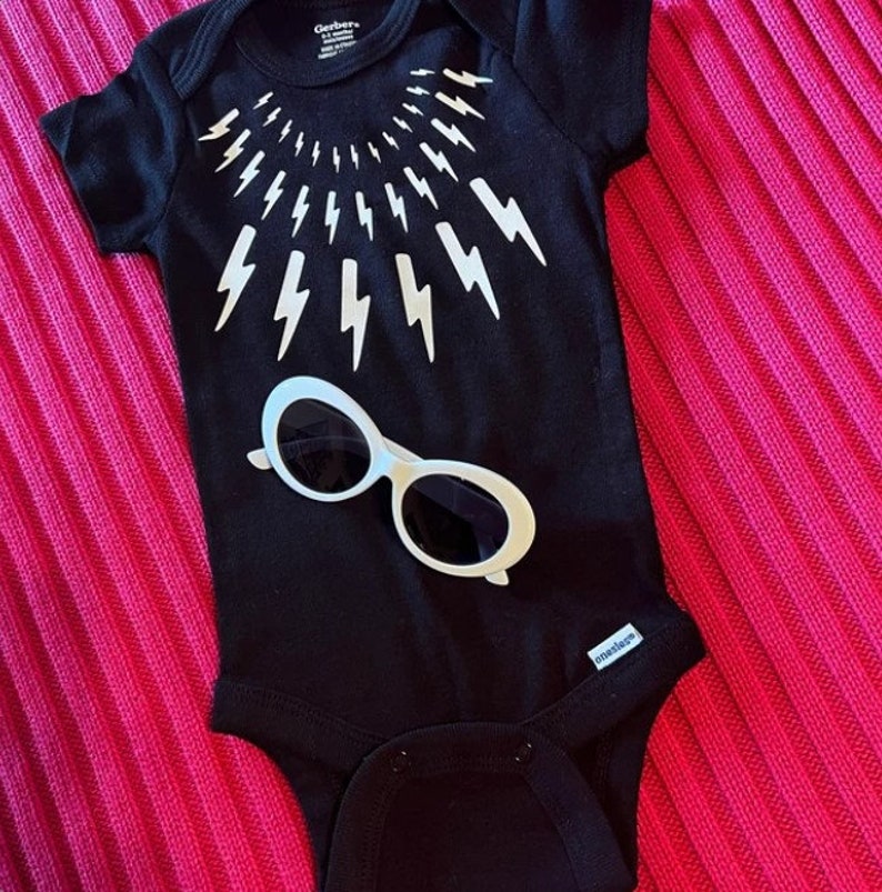 May include: Black baby onesie with a white lightning bolt design around the neck and a pair of white sunglasses on the front. The onesie has the Gerber brand name on the tag.