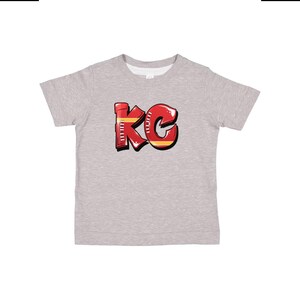 May include: A heather grey t-shirt with a red and yellow graphic of the letters "KC" in a graffiti style.