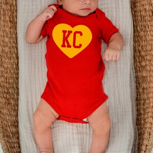 May include: A baby wearing a red onesie with a yellow heart that says "KC" on it. The baby is lying in a wicker basket.